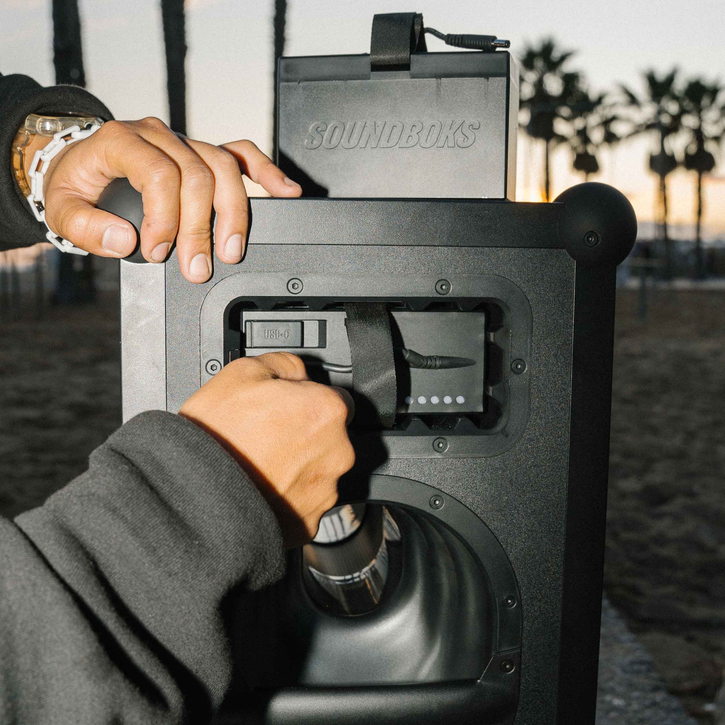 Close-up of a person inserting THE BATTERY into a black SOUNDBOKS 4 party speaker outdoors at sunset. The background features palm trees and a beach setting. The person is wearing a dark sweatshirt and a wristwatch, preparing for an evening of music by the shore.